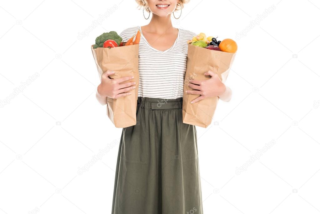 cropped shot of smiling young woman holding paper bags with fruits and vegetables isolated on white