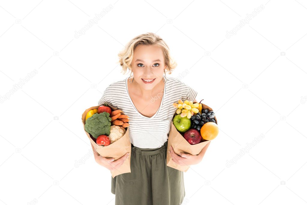 high angle view of beautiful young woman smiling at camera while standing with paper bags full of fresh fruits and vegetables isolated on white 