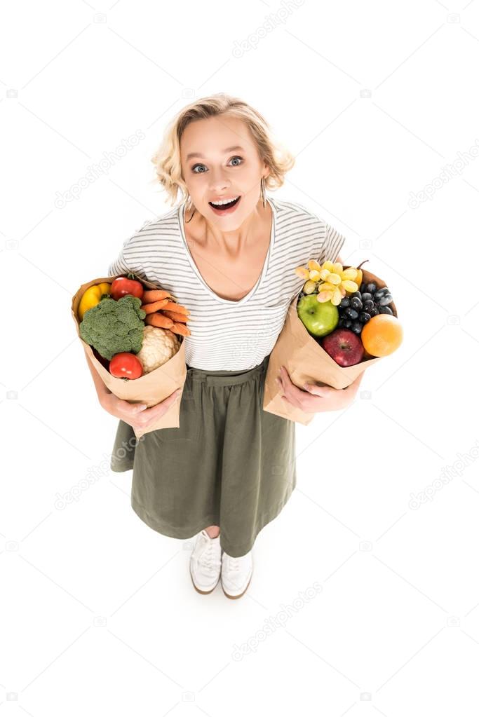 high angle view of cheerful young woman smiling at camera while standing with paper bags full of fresh fruits and vegetables isolated on white 