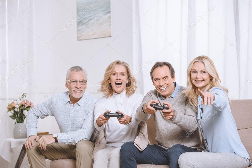 Middle aged men and women sitting on sofa and playing game console using joysticks
