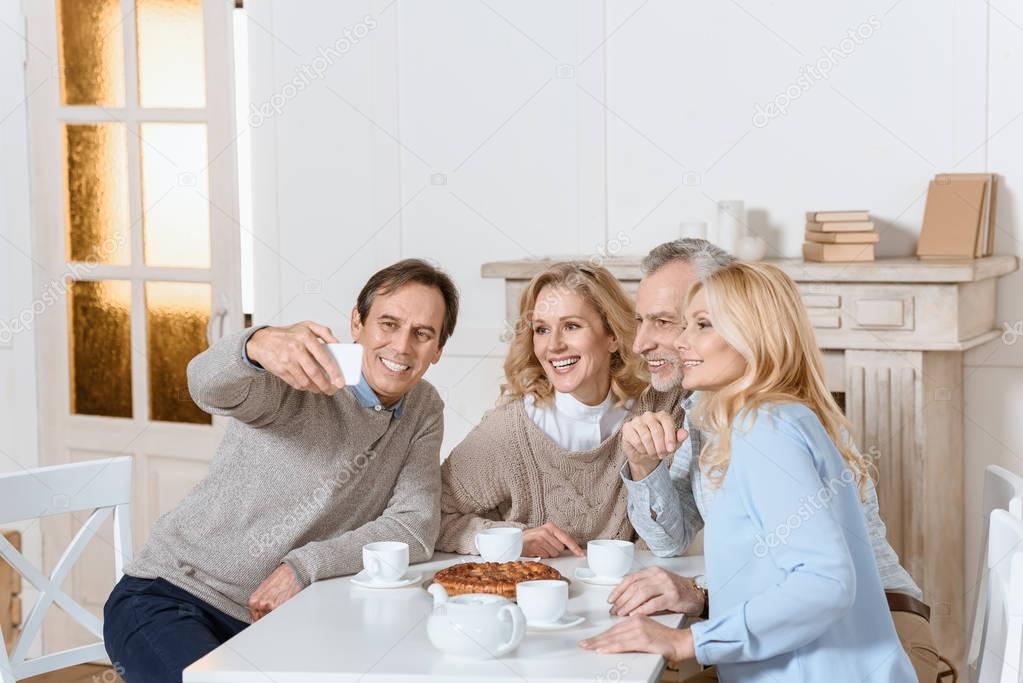 man doing selfie while friends sitting at table with tea and pie  