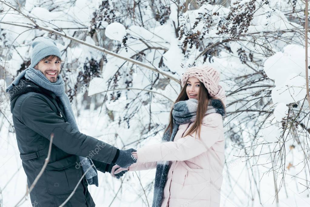 smiling couple holding hands and looking at camera in snowy forest