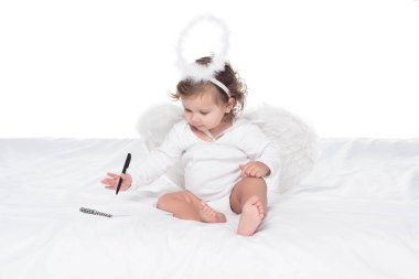 little angel with wings and nimbus writing in notepad on bed, isolated on white clipart