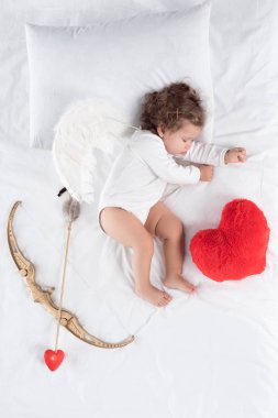 little baby with wings lying on bed with heart pillow, bow and arrow  clipart