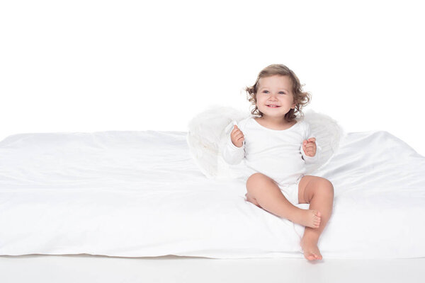 adorable happy baby with wings sitting on bed, isolated on white