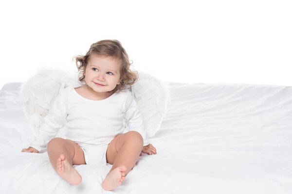 adorable smiling baby with wings sitting on bed, isolated on white
