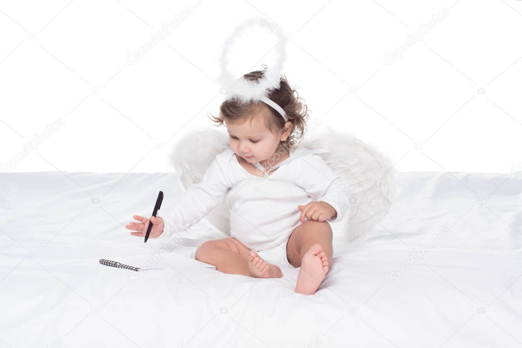 little angel with wings and nimbus writing in notepad on bed, isolated on white