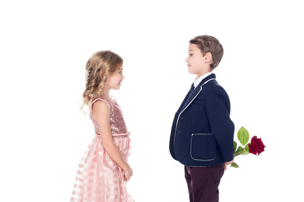 side view of fashionable boy in suit holding rose flower and looking at adorable little girl in pink dress isolated on white 