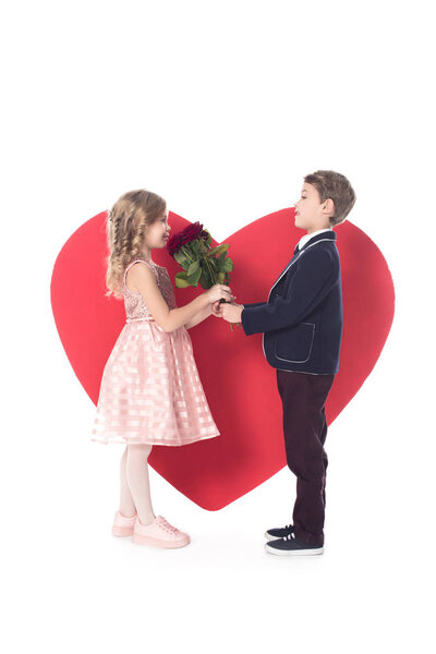 side view of cute little boy presenting flowers to adorable little girl and big red heart symbol behind isolated on white 