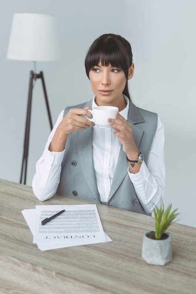 attractive businesswoman drinking coffee and looking away isolated on gray