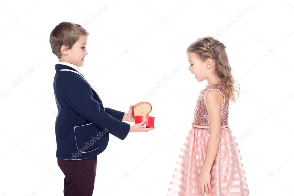 side view of stylish boy opening and presenting heart shaped gift box to surprised little girl isolated on white 