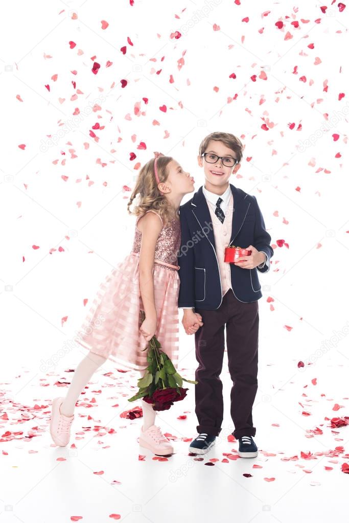 cute little boy in eyeglasses and suit holding gift box and smiling at camera while girl with roses able to kiss him on white with falling heart shaped confetti