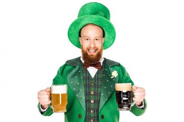 cheerful bearded man in green costume and hat holding glasses of beer and looking at camera isolated on white  
