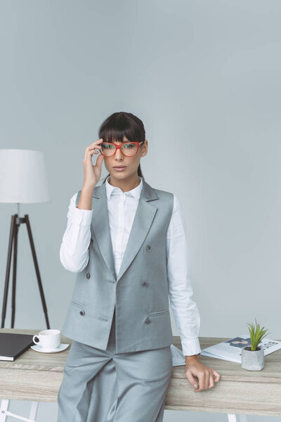 businesswoman touching glasses and looking at camera isolated on gray