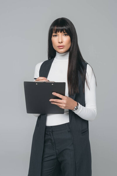 businesswoman standing with clipboard and looking at camera isolated on gray