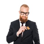 Homme d'affaires barbu souriant mettant la carte de crédit dans la poche de la veste de costume isolé sur blanc