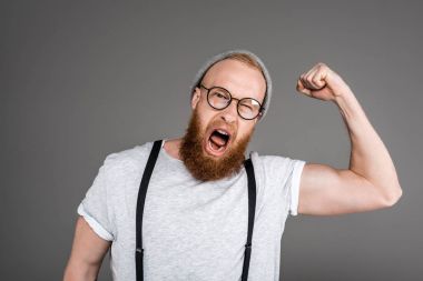 excited bearded man showing biceps and screaming at camera isolated on grey clipart