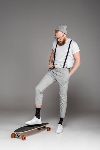stylish bearded man standing on longboard and looking down on grey