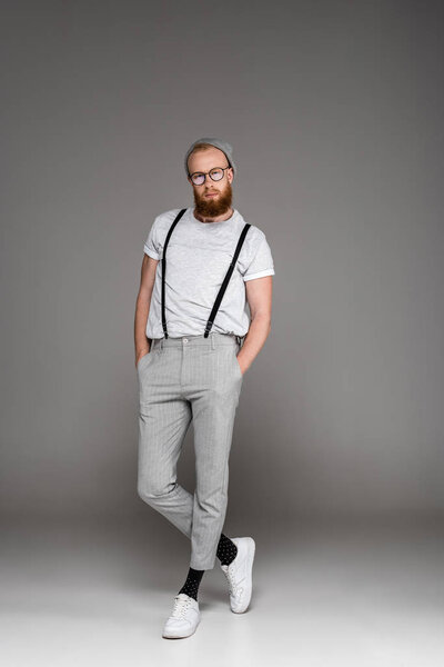 стильный бородатый мужчина в шляпе и очках, стоящий с руками в карманах и смотрящий в камеру на сером
