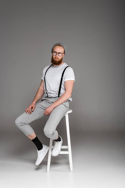 стильный бородатый хипстер в глазницах сидит на стуле и смотрит в камеру на сером
  