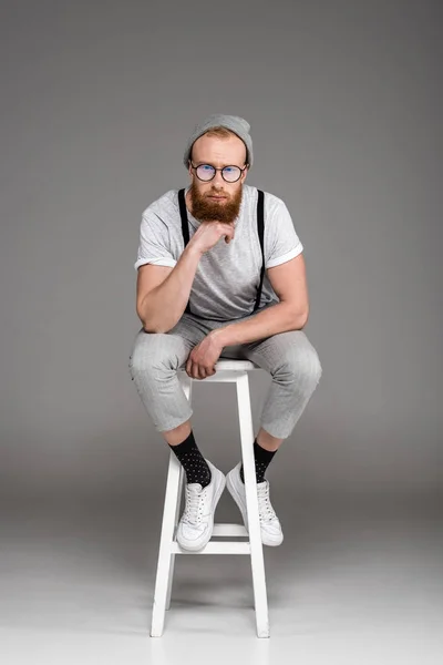 stylish bearded man in eyeglasees sitting on stool and looking at camera on grey