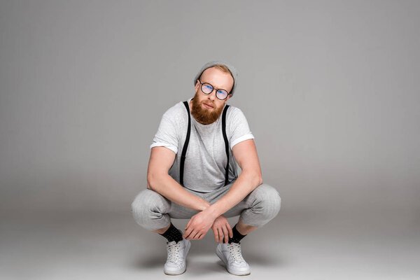 stylish man in hat and eyeglasses crouching and looking at camera on grey 