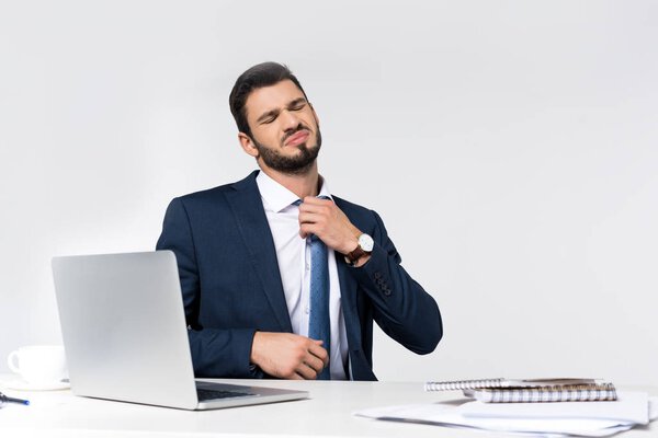 young overworked businessman with closed eyes adjusting necktie while sitting at workplace 