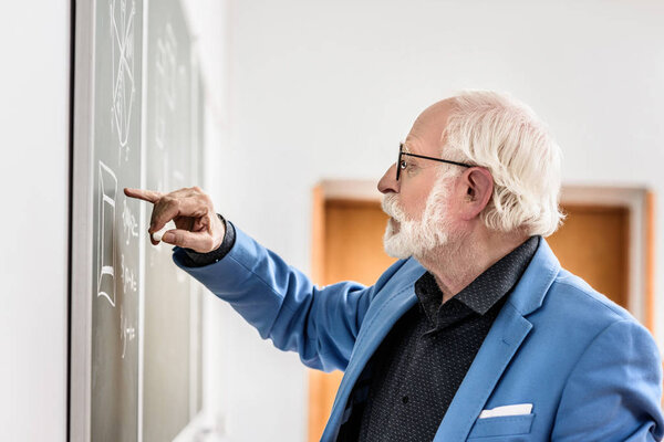 side view of grey hair professor correcting something at blackboard