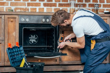young repairman in protective workwear fixing oven with electric drill clipart