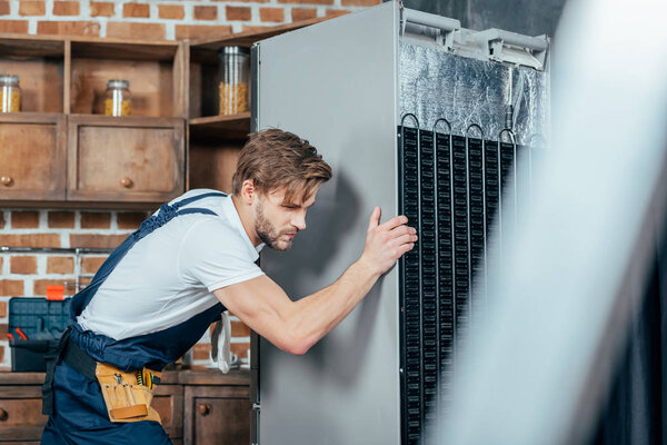 young repairman in protective workwear moving refrigerator in kitchen