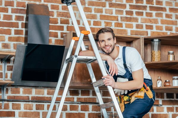 handsome young foreman standing on ladder and smiling at camera while fixing kitchen hood 