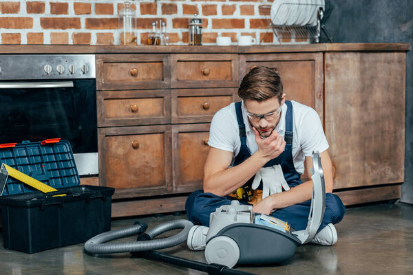 young repairman in protective glasses looking at broken vacuum cleaner 