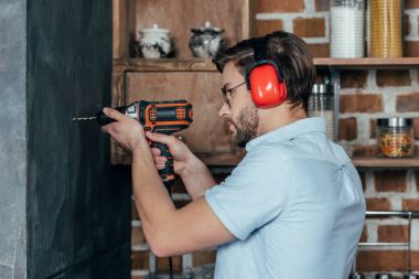 side view of young man in eyeglasses and earmuffs drilling wall with electric drill clipart