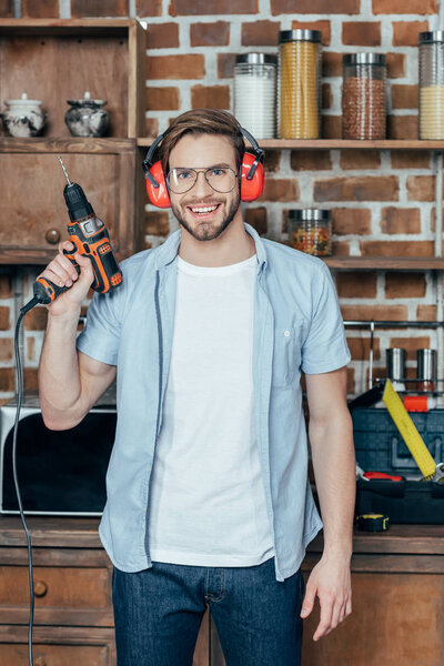 handsome young man in eyeglasses and earmuffs holding electric drill and smiling at camera