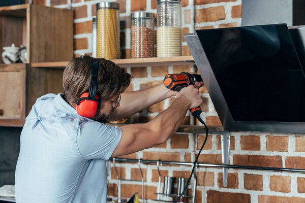 young man in earmuffs drilling kitchen hood with electric drill