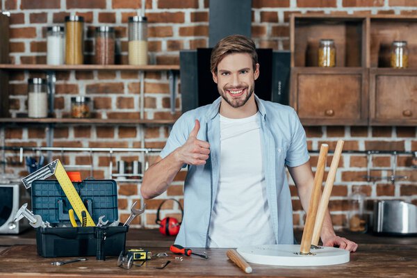 handsome young man smiling at camera and showing thumb up while repairing chair at home