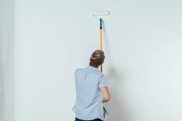 back view of young man using paint roller while painting wall at home