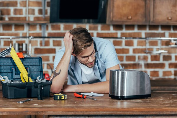 frustrated young man in eyeglasses looking at broken toaster at home