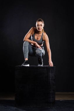 athletic young sportswoman crouching on black cube and looking at camera on black clipart