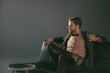young businessman holding glass of whiskey and looking away isolated on grey clipart