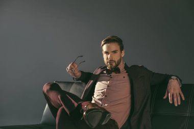 gözlük tutan ve kanepede otururken kameraya bakarak şık yakışıklı genç