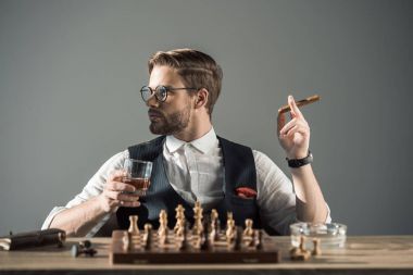 young man with glass of whisky smoking cigar and looking away while playing chess  clipart