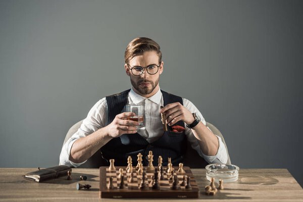 young businessman with glass of whisky and cigar looking at camera while playing chess