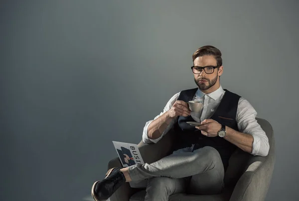 コーヒーを飲むと 灰色に分離された肘掛け椅子で新聞と一緒に座ってカメラ目線の眼鏡でハンサムな青年実業家 — ストック写真