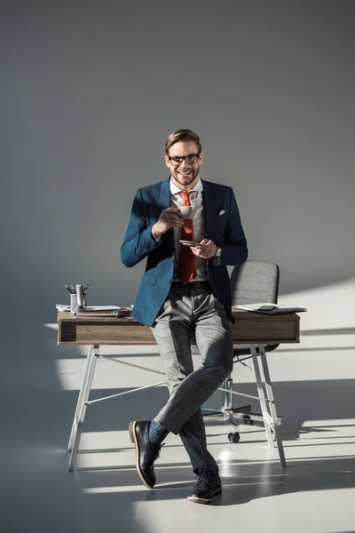 一杯のコーヒー テーブルの上に座って グレーにカメラ目線で笑顔のスタイリッシュなビジネスマン — ストック写真