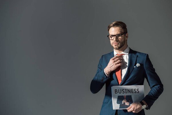 handsome young man in eyeglasses holding newspaper and adjusting necktie isolated on grey 