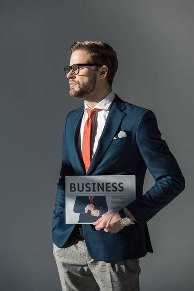 handsome young businessman holding newspaper and looking away isolated on grey