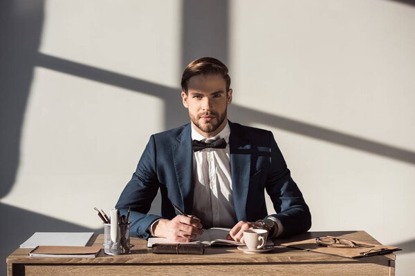 handsome stylish businessman looking at camera while writing in diary at workplace