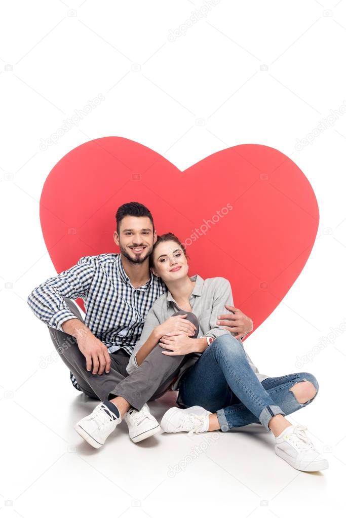 happy couple sitting near big heart on white, valentines day concept