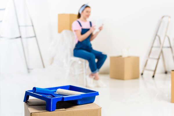 girl sitting with packed boxes in new house with paint tray on foreground
